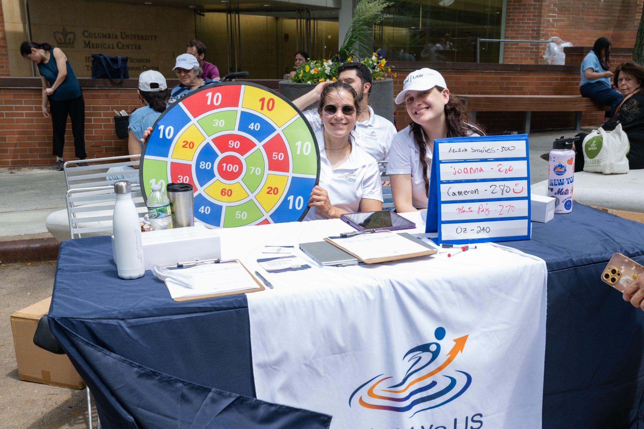 Two Let's Move US volunteers smiling at the booth table, with the Columbia University Medical Center building behind them