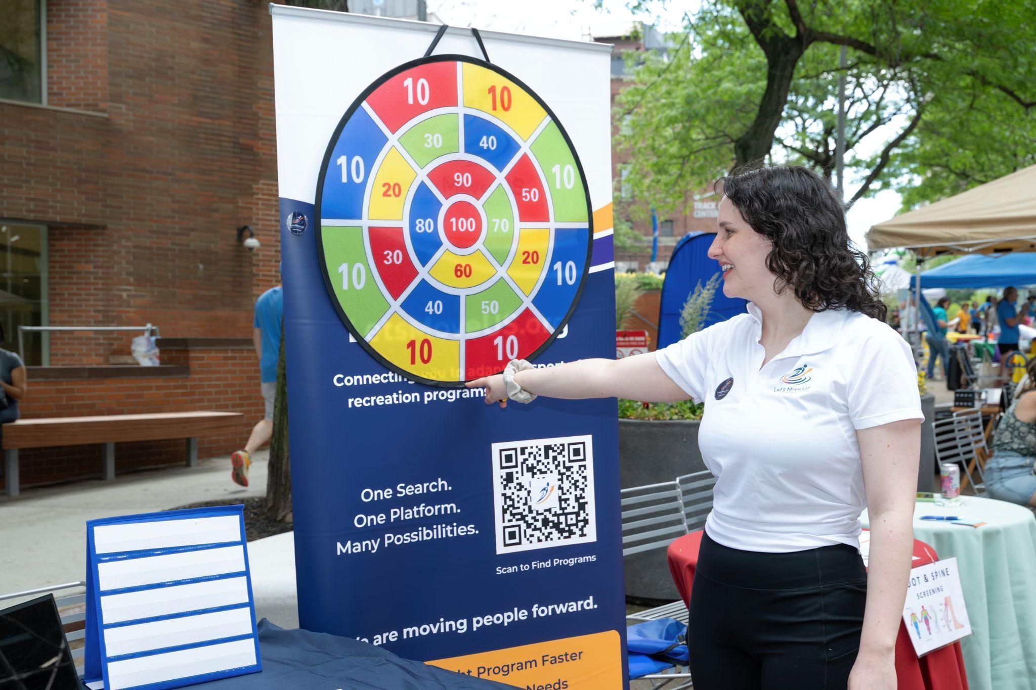 A volunteer in a Let's Move US polo pointing at a banner featuring a dartboard game and a QR code reading 'One Search. One Platform. Many Possibilities.'